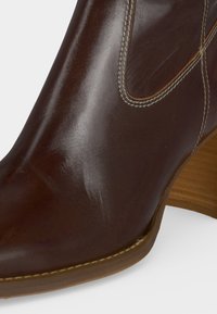 Bottine en cuir marron avec panneau élastique latéral, talon empilé en bois et coutures visibles le long de la semelle et des bords du panneau.