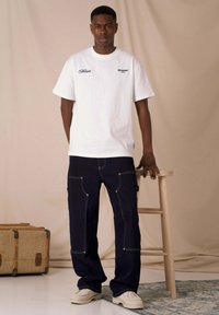 Camiseta blanca de algodón con el logo "SKYTHUNE PARIS", combinada con pantalones de trabajo azul oscuro con costuras visibles y bolsillos laterales, y zapatos color crema.