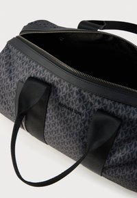 Μαύρη τσάντα duffle με σχέδιο λογότυπου MK σε γκρι χρώμα, λείο ύφασμα, μαύρες δερμάτινες λεπτομέρειες και κλείσιμο με φερμουάρ. Εσωτερική άποψη του χώρου αποθήκευσης.