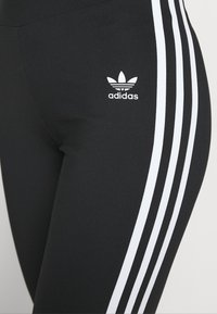 Zwarte leggings met een witte drie-strepen ontwerp langs de zijkanten en een klein wit adidas-logo op de linker bovenbeen. Glad textiel.
