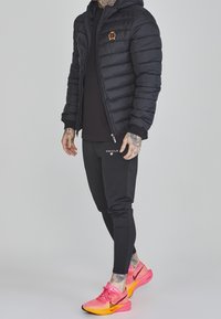 Černá péřová bunda s kapucí, výšivkou emblému, v kombinaci s černými tepláky a jasně růžovými teniskami Nike s oranžovými akcenty.