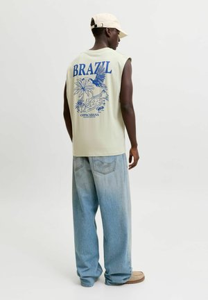 Homme portant un débardeur avec un imprimé bleu Brésil et Copacabana, un jean large bleu clair, une casquette beige et des chaussures beiges, debout de côté.