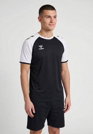 Mężczyzna w czarno-białej sportowej koszulce i czarnych szortach, stojący i lekko patrzący w prawo, na jednolitym jasnoszarym tle.