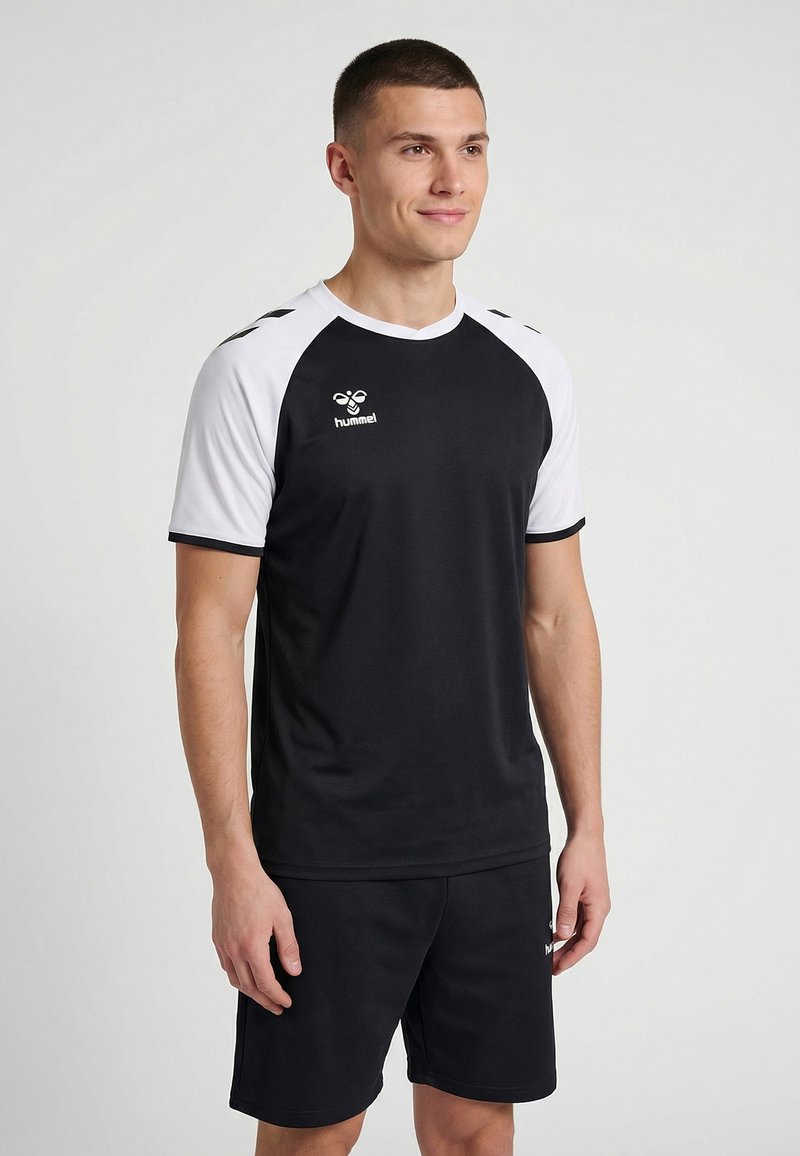 Man in zwart-wit sportshirt en zwarte korte broek, staand en iets naar rechts kijkend, tegen een effen lichtgrijze achtergrond.