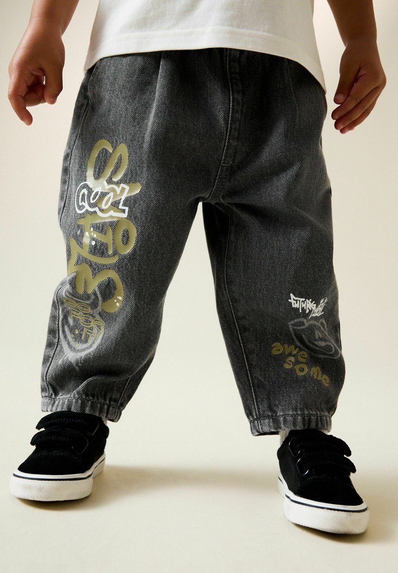 Dunkelgraue Jeanshose mit lockerem Schnitt, versehen mit weißen und metallic-goldenen Grafikdrucken und einem elastischen Bund. Schwarze Turnschuhe vervollständigen das Outfit.