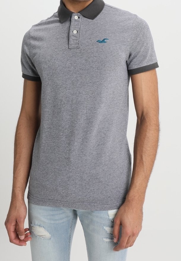 Polo gris avec un col noir contrastant, à manches courtes, et un logo turquoise sur la poitrine, associé à un jean léger à déchirures.