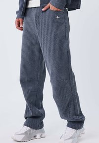 Pantalon à carreaux bleu marine en tissu texturé, doté d'une poche latérale et d'un marquage subtil. Associé à des chaussures de sport blanches.