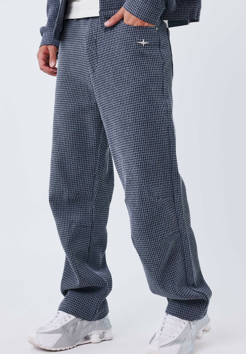 Pantalon à carreaux bleu marine en tissu texturé, doté d'une poche latérale et d'un marquage subtil. Associé à des chaussures de sport blanches.