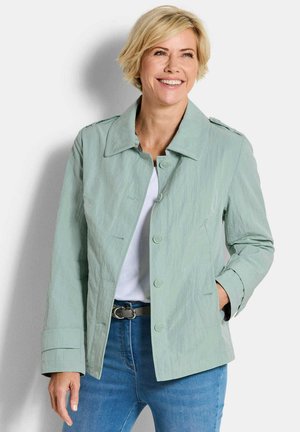 Lächelnde Frau mittleren Alters mit kurzen blonden Haaren, die eine hellgrüne, geknöpfte Jacke, ein weißes Hemd, blaue Jeans und einen schwarzen Gürtel trägt.