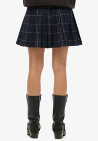 Jupe plissée bleu marine à motif à carreaux, avec des lignes bleu clair. Bottes en cuir noires montantes avec des boucles.