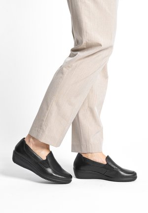 Person wearing beige pants and black slip-on wedge shoes, one foot slightly raised as if walking, set against a white background.