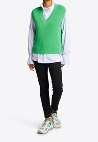 Person, die einen grünen ärmellosen Pullover über einer gestreiften, rüschengesäumten Bluse trägt, schwarze schmale Hosen und weiße Sneakers mit grünen Akzenten.