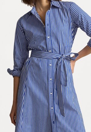 Femme portant une robe chemise rayée verticale bleue et blanche avec une ceinture nouée à la taille et des manches retroussées.