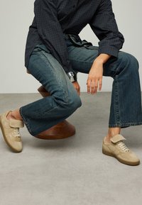 Beige sneakers with a suede upper and rubber sole, featuring a flap overlay. Paired with dark denim jeans and a checkered shirt.
