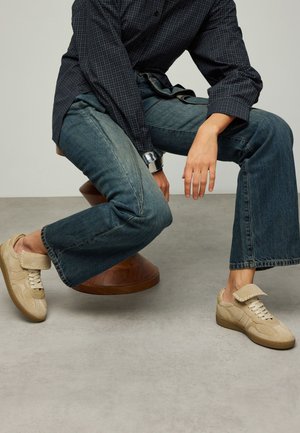 Beige Sneaker mit einem Obermaterial aus Wildleder und einer Gummisohle, mit einer Überlappung aus Klappe. Kombiniert mit dunklen Denim-Jeans und einem karierten Hemd.