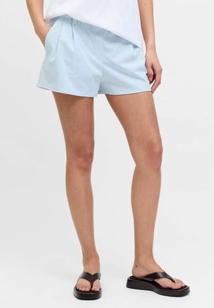 Kvinde iført lyseblå plisserede shorts, hvid top og sorte platformsflipflops, stående med den ene hånd i lommen på ensfarvet baggrund.