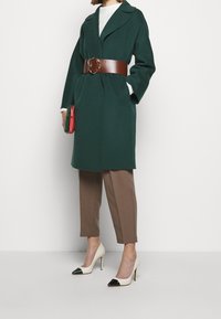 Manteau en laine vert avec taille ceinturée marron, associé à un pantalon beige et des talons crème à bouts noirs. Tient une pochette multicolore.