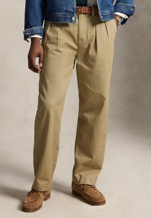 Homme portant un pantalon plissé beige, une ceinture tressée marron, une veste en jean bleue et des chaussures en daim marron, debout sur un fond clair.