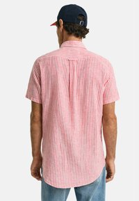 Camicia a maniche corte a righe verticali rosse e bianche, con un orlo arrotondato e una piega posteriore per un maggiore movimento. Senza loghi o decorazioni visibili.