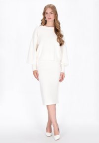 Biała dziana crop sweter z szerokimi rękawami w zestawie z dopasowaną białą spódnicą ołówkową. Modelka ma również na sobie białe czółenka z ostrym noskiem.