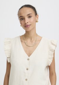 Frau mit zurückgebundenen lockigen Haaren, trägt goldene Creolen, eine goldene Halskette und eine elfenbeinfarbene ärmellose Bluse mit Raffungen an den Schultern und Knöpfen.