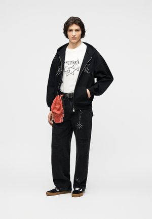 Persona joven con una sudadera negra con cremallera, camiseta blanca con gráficos, jeans negros con tachuelas, zapatillas negras y sosteniendo una gorra roja a un lado.