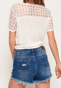 T-shirt bianca con maniche e schiena in pizzo, abbinata a pantaloni corti in denim blu con orlo sfrangiato e tasche logorate.