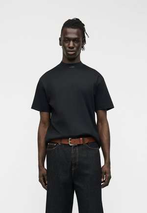 Uomo con maglietta nera a maniche corte con logo "BOSS", jeans scuri e cintura marrone, in piedi contro uno sfondo chiaro e uniforme.