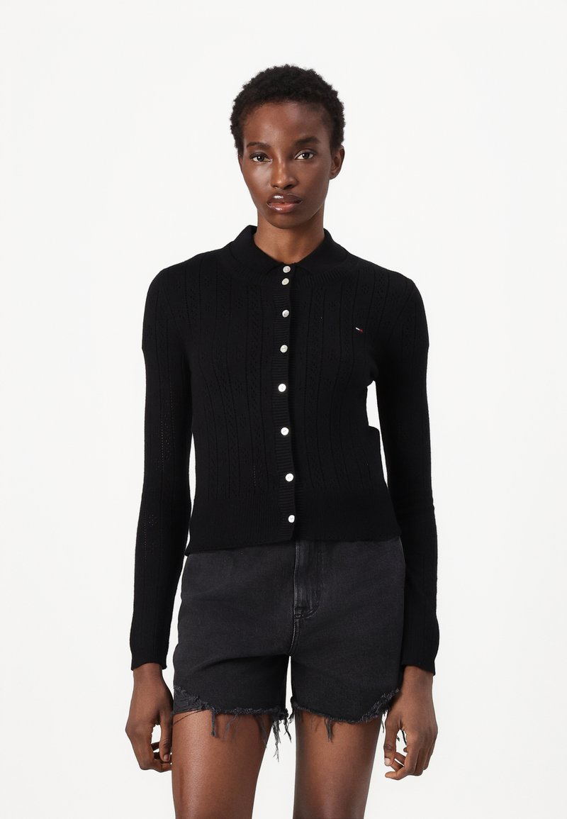 Svart cardigan med strukturert strikkedesign, ribbestrikket krage og knappåpning. Har elfenbensfargede knapper og minimal logo-detalj.
