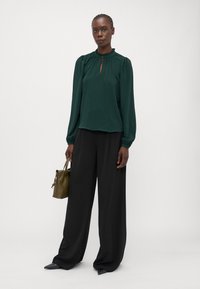 Groene blouse met lange mouwen, geplooide halslijn en sleutelgatdetail, gecombineerd met zwarte broek met wijde pijpen en een kleine olijfgroene handtas.