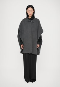 Sivi vuneni ogrtač s visokim ovratnikom, pričvršćen crnim detaljima, nošen preko tamne bluze i crnih širokih hlača. Glatka tekstura i suvremen dizajn.