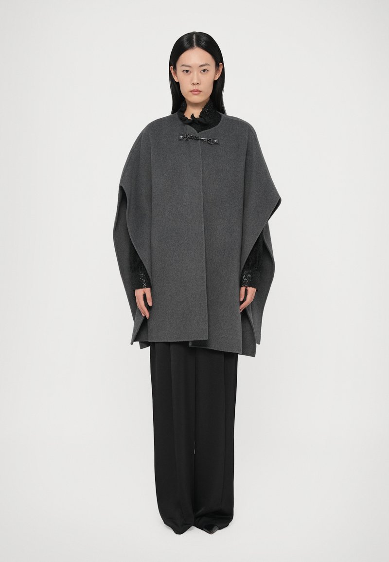 Sivi vuneni ogrtač s visokim ovratnikom, pričvršćen crnim detaljima, nošen preko tamne bluze i crnih širokih hlača. Glatka tekstura i suvremen dizajn.