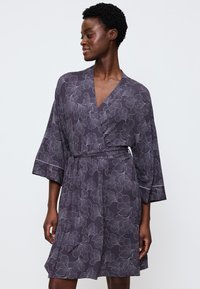 Donna che indossa un vestito a portafoglio floreale viola scuro, lungo fino al ginocchio, con maniche ampie, mentre guarda a sinistra su uno sfondo chiaro e semplice.