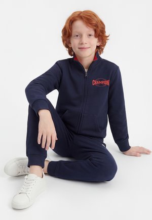 Ung rødhåret dreng sidder med benene over kors iført en marineblå træningsdragt med lynlås og "Champion"-logo samt hvide sneakers på hvid baggrund.