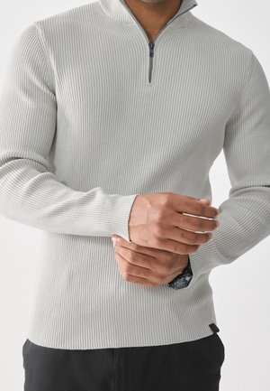 Homme portant un pull côtelé gris clair avec fermeture éclair au quart, pantalon noir et une montre noire au poignet, ajustant la manche avec ses mains.