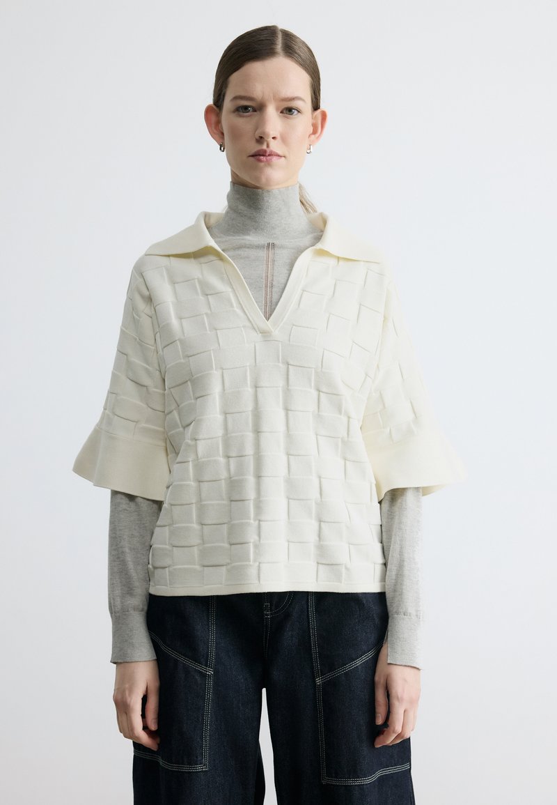 Femme portant un haut blanc texturé à manches courtes superposé à un col roulé gris et un pantalon large foncé avec des coutures blanches.