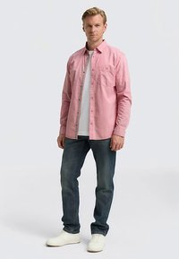 Camicia rosa con bottoni e taschino, indossata sopra una t-shirt bianca, abbinata a jeans blu scuro e sneakers bianche. Tessuto strutturato con colletto.