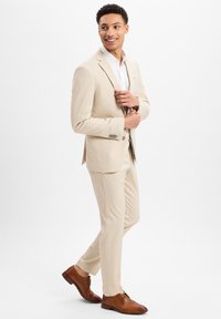 Lichtbeige pak met een getailleerde pasvorm, een enkelzijdige blazer en bijpassende broek. Witte blouse eronder en bruine leren schoenen.