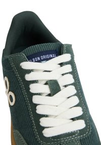 Zapatilla de pana verde con cordones blancos, que cuenta con un cuello acolchado y un detalle de logo. Incluye una etiqueta azul en la lengüeta.