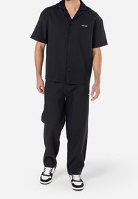 Camicia nera a maniche corte con bottoni, dal tessuto a coste, dotata di colletto e logo sul petto, abbinata a pantaloni neri loose-fit e sneakers.