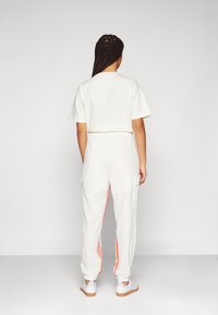 Henkilö, jolla on pitkät letitetyt hiukset, pukeutuneena valkoiseen lyhythihaiseen paitaan, valkoisiin housuihin, joissa on oranssit sivuraidat, sekä valkoisiin lenkkareihin, selkä kameraan päin yksinkertaisella taustalla.