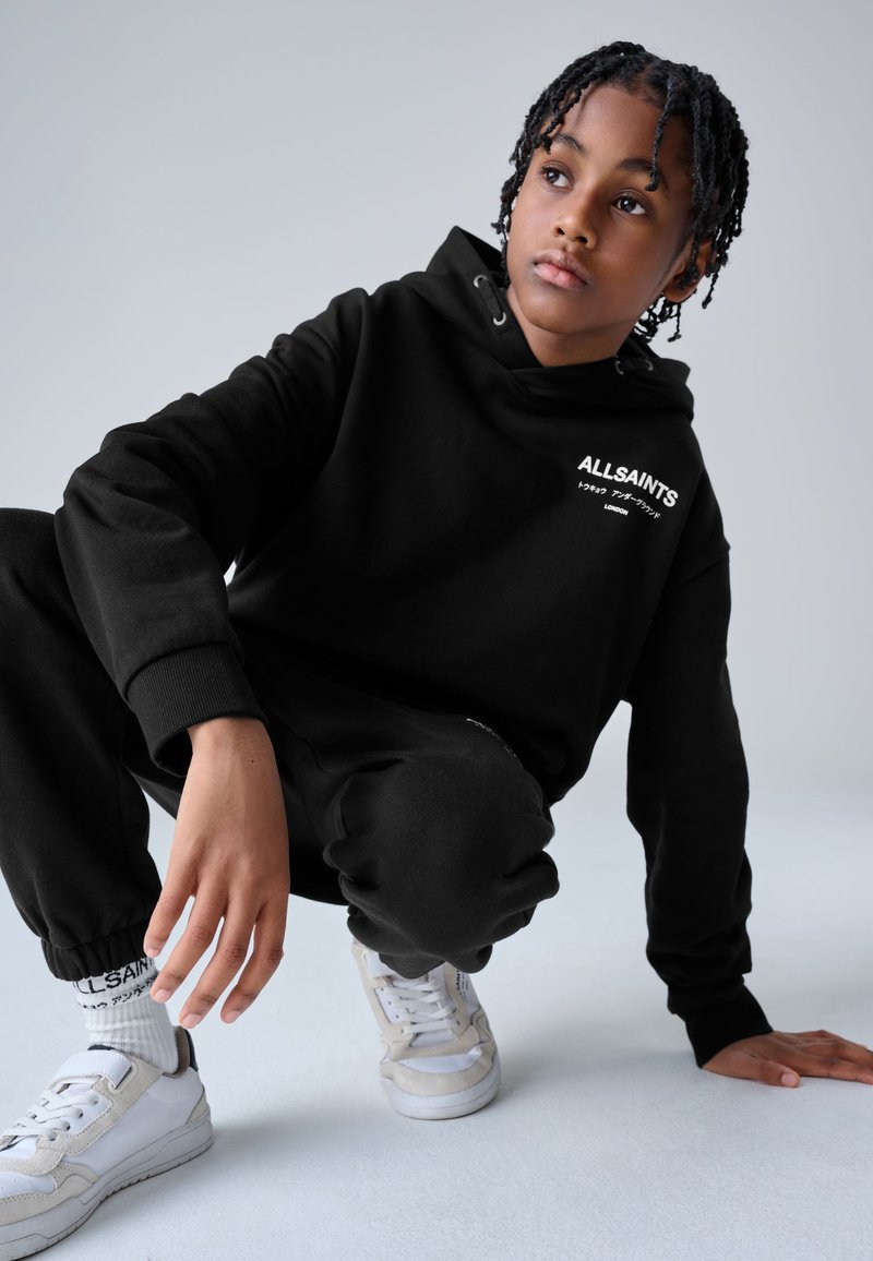 Giovane con capelli intrecciati che indossa una felpa nera AllSaints con cappuccio, pantaloni neri, sneakers bianche e calzini bianchi, accovacciata su uno sfondo chiaro.