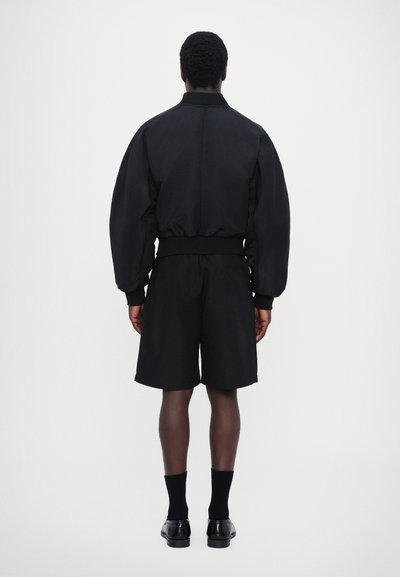 Veste bomber noire avec une texture lisse, des poignets élastiques, associée à un short noir. La tenue a un design minimaliste.