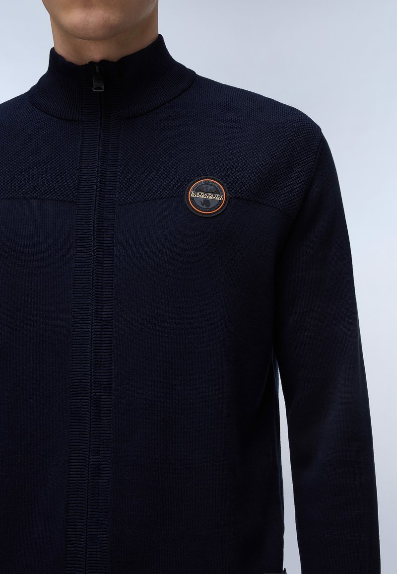 Pull en maille bleu marine zippé avec un col montant, des panneaux texturés et un badge logo rond sur la poitrine. Fabriqué en tissu tricoté.