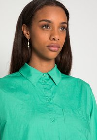 Camisa verde de botão, feita de tecido leve, com um colarinho clássico, bolso frontal e textura suave.