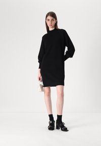 Fekete kötött, hosszú ujjú ruha magas nyakkivágással, fekete zoknikkal és vaskos sarkú szandálokkal, amelyek arany részletekkel rendelkeznek. Minimalista kézitáska.