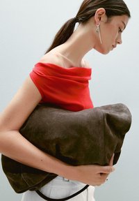 Suede brown shoulder bag held against a red off-shoulder top. The bag features a soft texture and rounded shape, with minimalistic design accents.
