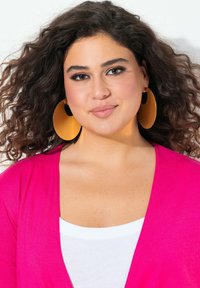 Cheveux bouclés, portant de grandes boucles d'oreilles en or rondes. Habillée d'un cardigan rose par-dessus un haut blanc. Fond neutre.