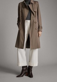 Personne portant un trench-coat marron croisé, un pull à col roulé gris, un pantalon blanc à jambes larges et des bottines marron foncé.