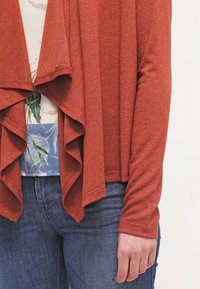 Rdzawy kardigan z długim przodem, miękką teksturą i długimi rękawami, nałożony na kwiecistą bluzkę i zestawiony z niebieskimi dżinsami.